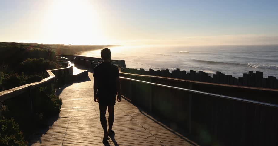 A man looks at the ocean as he walks down a wood plank path to the beach