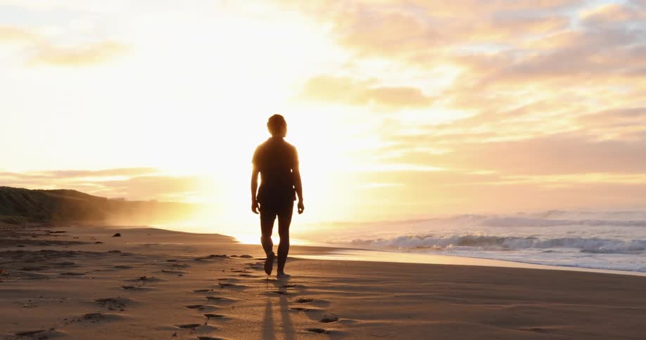 The setting sun glows around a man walking barefoot on the beach as the ocean waves curl and splash