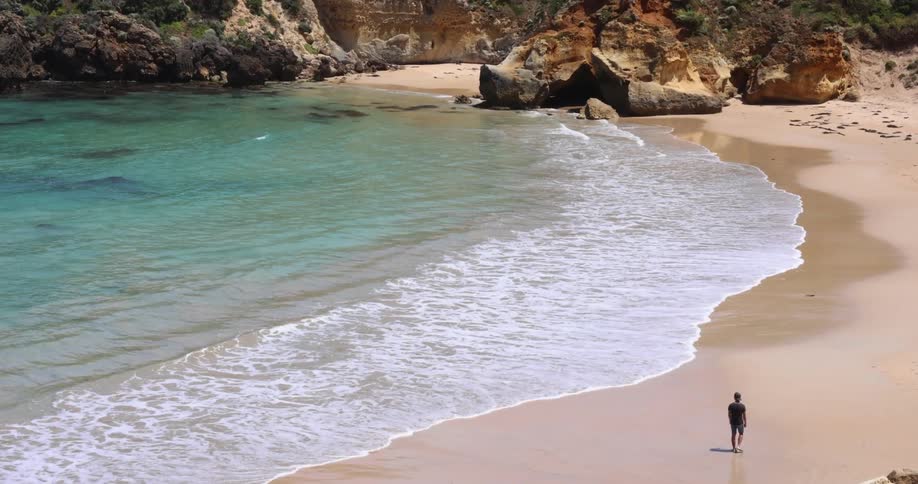 A person walking on a beach with sand and waves with a rocky cliff in the background