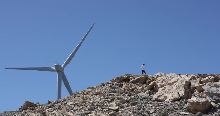 A man walks near a wind turbine at the top of a rocky hill