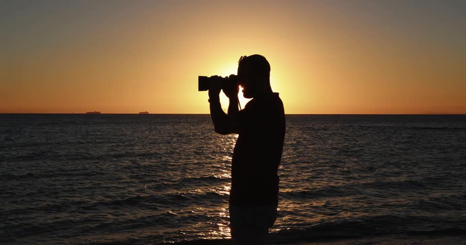 A silhouette of a man with a camera on a beach with the ocean the sunset the sky and clouds in the background