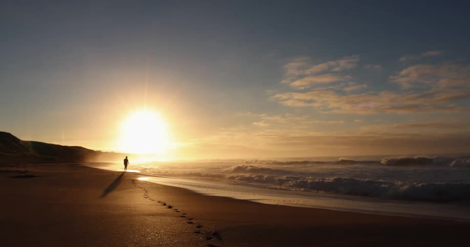 Person stands and walks along beach leaving footprints as sun sits on horizon
