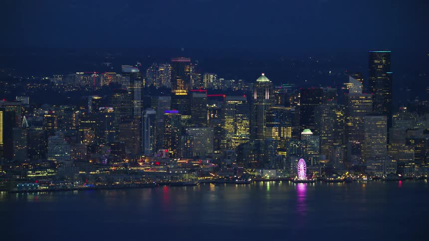 Wide aerial of Downtown Seattle looking east across the bay to the sky scrapers beyond as they light up in night lights