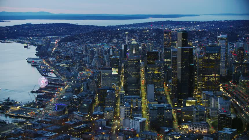 The Seattle Great Wheel glows pink on the waterfront as the viewer looks straight down city streets and a busy highway with skyscrapers rising above the city of Seattle