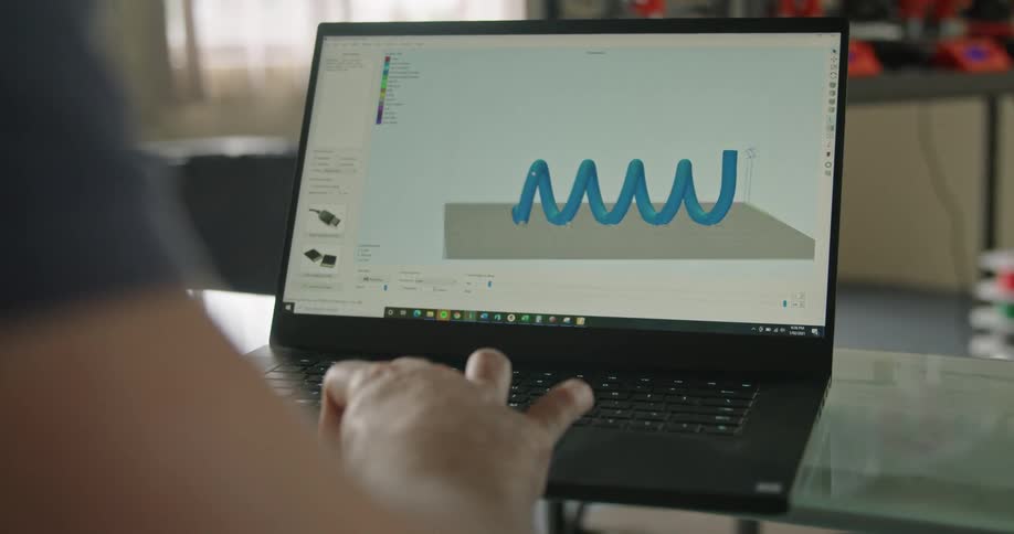 A person using a laptop with a 3d printer a wall and window in the background