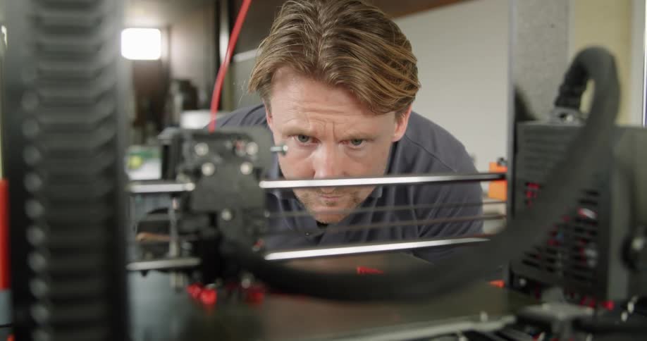 A man looking at a 3d printer printing with a wall and a door in the background