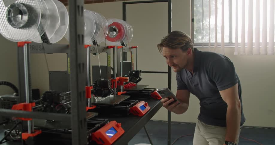 A man takes pictures with his smartphone of a table with 3d printers