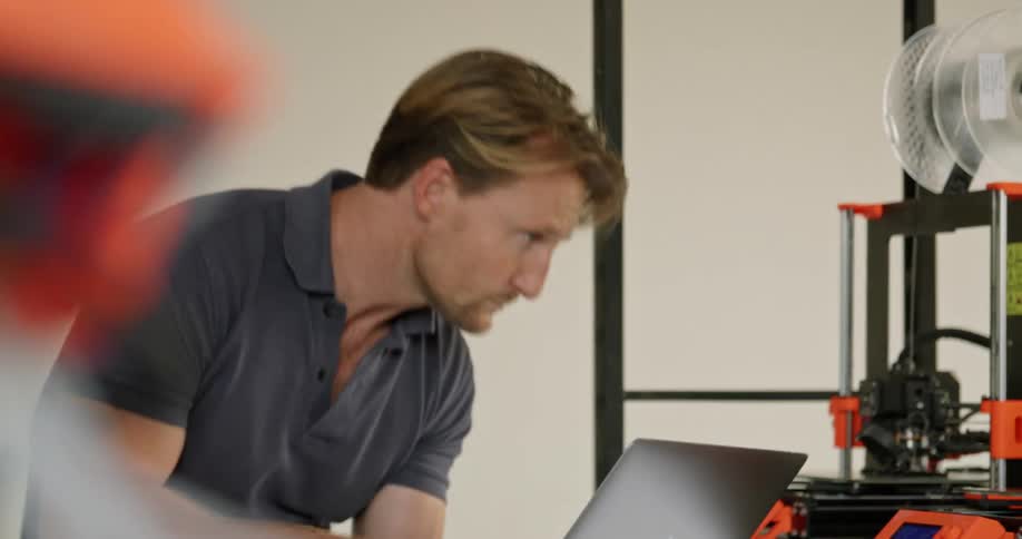A man types on his laptop as he monitors and watches 3d printers printing