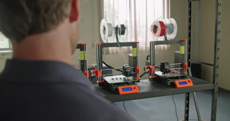 A man stands in a room watching three 3d printers printing