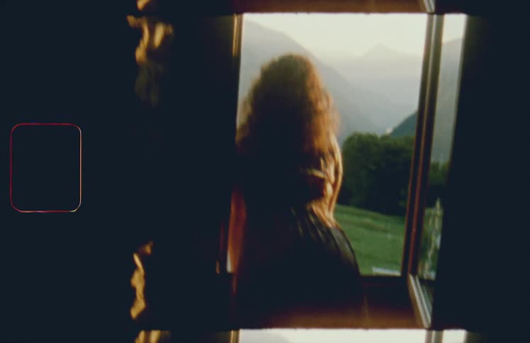 A woman opens a window and leans out looking at hills and a large green lawn