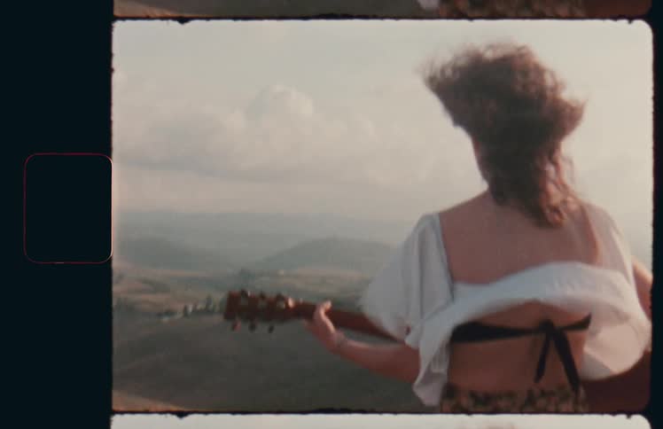 A woman with a guitar standing in fierce wind at high altitude looking out at the countryside below as her clothing whips around her