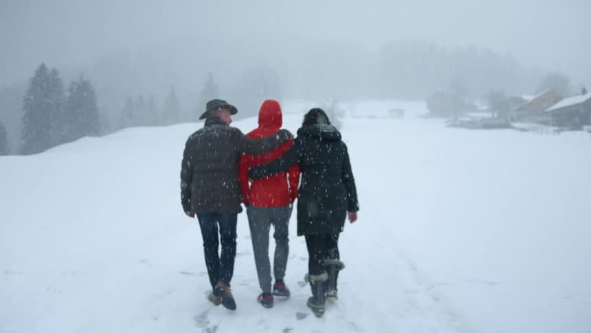 A man and woman embrace a third person from each side while walking through the snowy weather on a foggy day