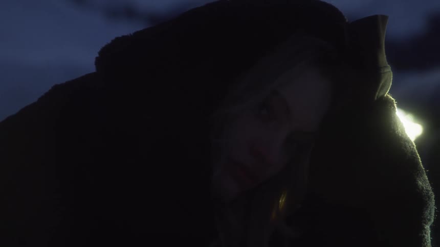 A woman wearing a hooded sweater leans forward and presses her sleeve to her head while she is outdoors at night