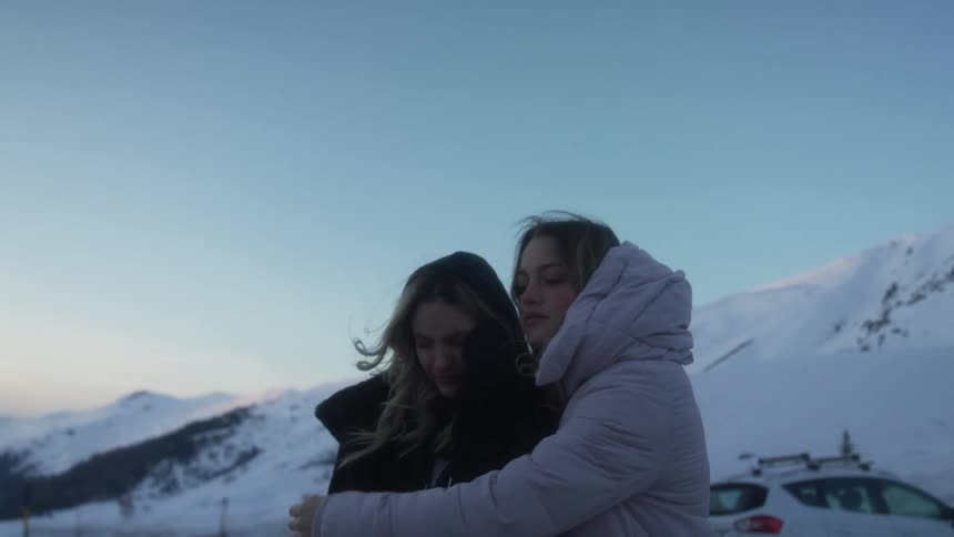 Two young women embrace as they stand near their vehicle parked below a snowy mountain range