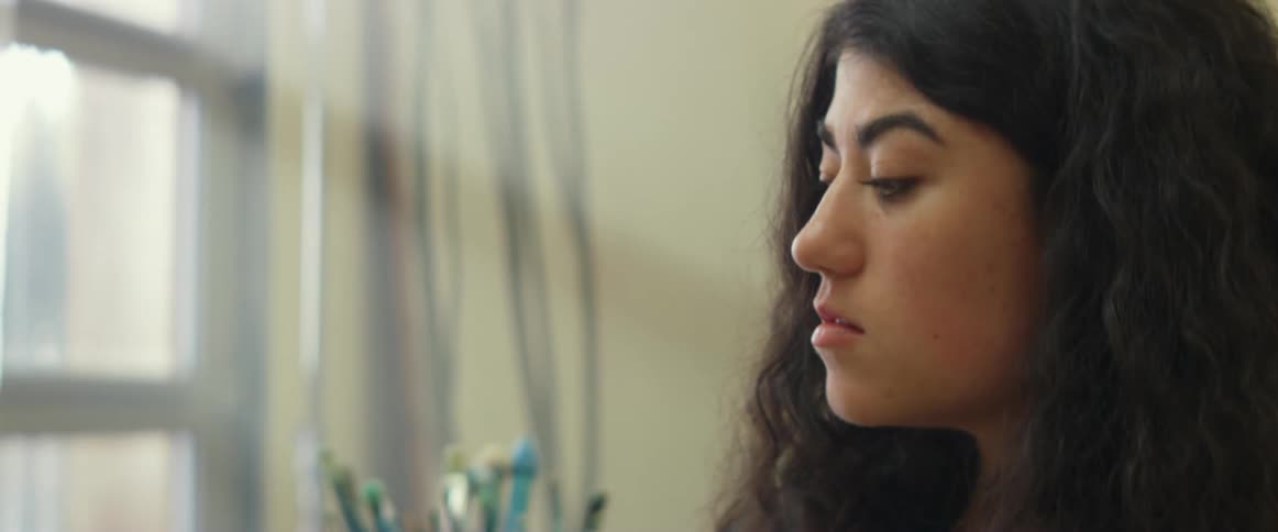 A woman in a room painting on a canvas with a paint brush with a wall and window in the background