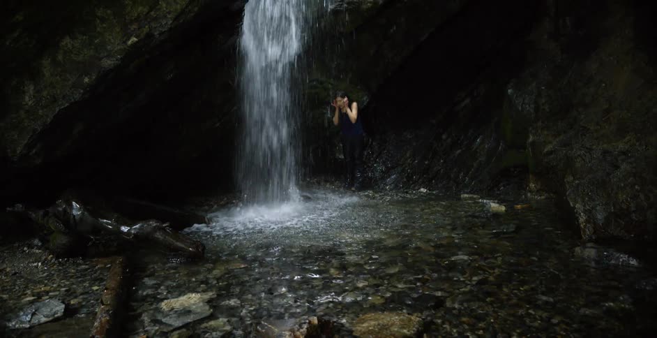 A young woman stands by a waterfall and splashes water on her face