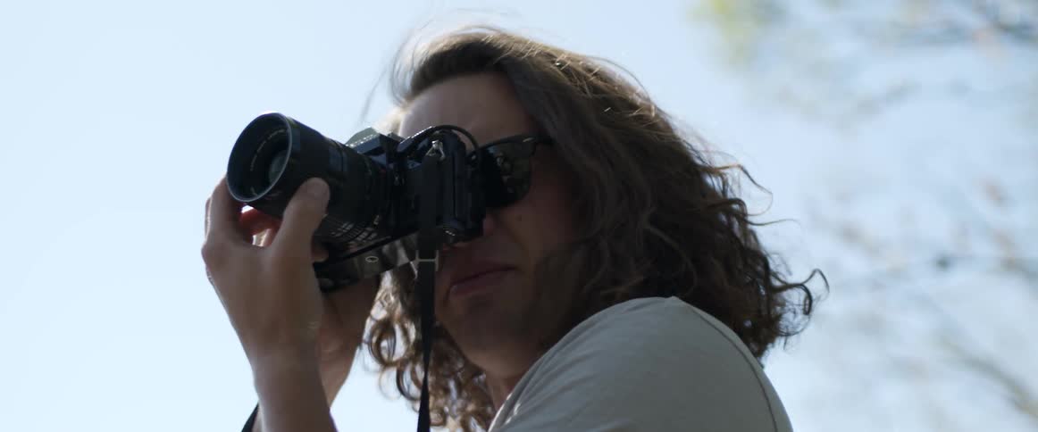 A man with sunglasses and a camera with the sky and clouds in the background