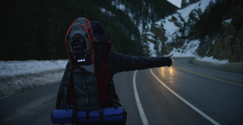 Young woman carrying a backpack hitchhiking on a road in the snow covered mountains