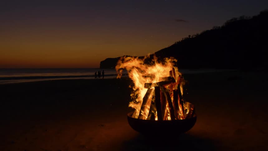 A large bonfire burns on the beach as people walk by the water