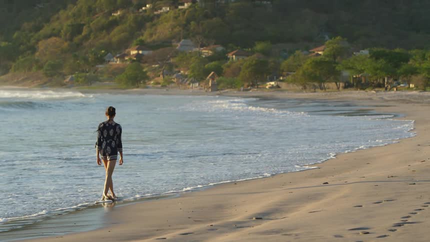 Woman walks along beach toward town sitting on forest covered mountain slope