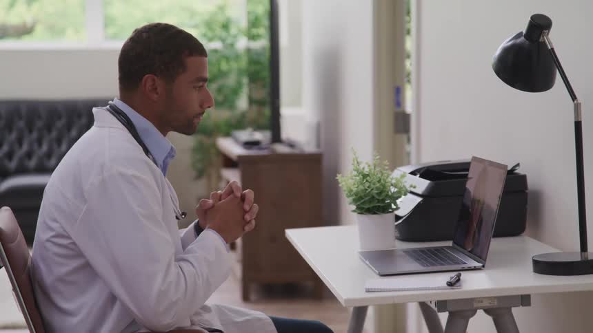 Male doctor with a white lab coat sits at desk and makes a video call on a laptop in the clinic