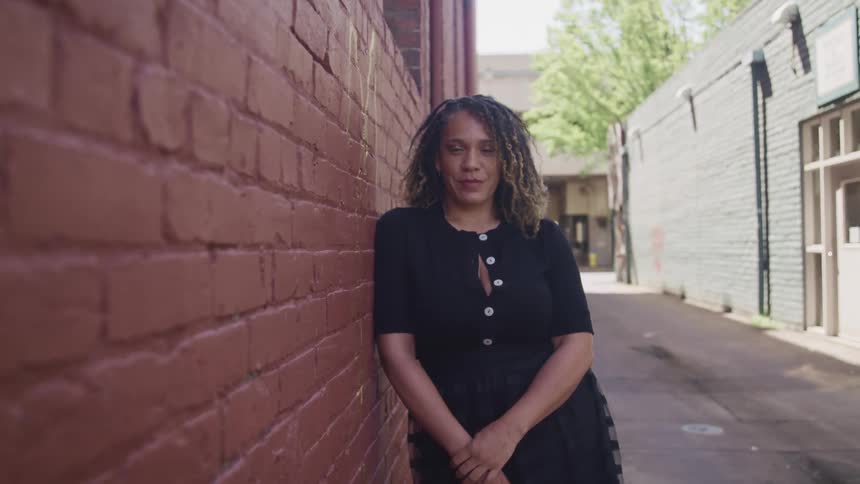 A woman leans and poses against a red brick building in an alley