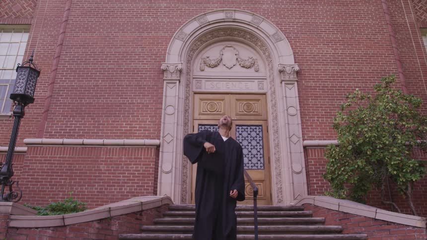 A college graduate throwing their graduation cap into the air on the steps of a university building