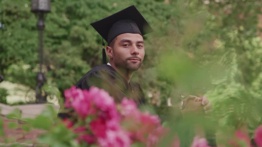Man wearing graduation gown sits in park near pink flowers