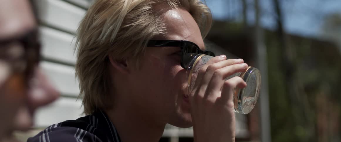A man with sunglasses drinking from a glass with a house trees the sky and clouds in the background