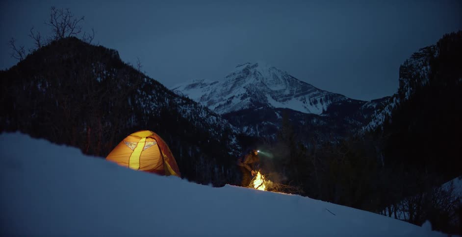 A person standing next to a glowing tent and fire while camping in snowy mountains at night