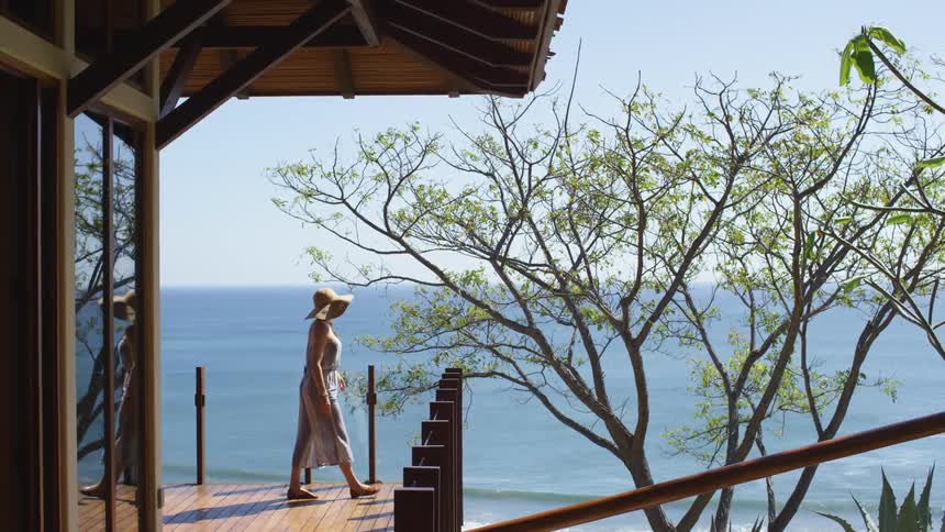 A woman with a hat walking on a deck with trees the ocean the sky and clouds in the background