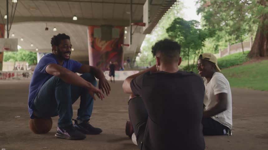 Three male friends smiling and laughing while sitting on an outdoor basketball court