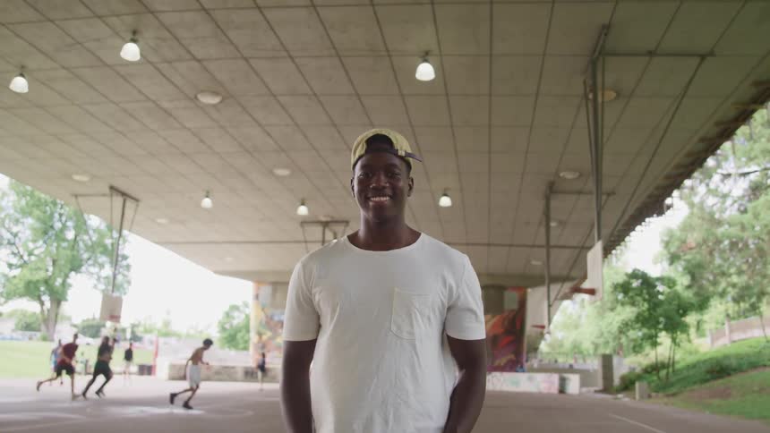Basketball player with a white t shirt and a cap faces the camera with eye contact in the basketball court under the bridge