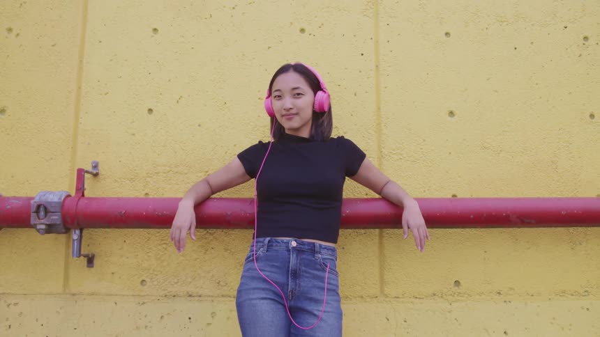 Young woman wearing pink headphones reclined on a red pipe on a yellow wall