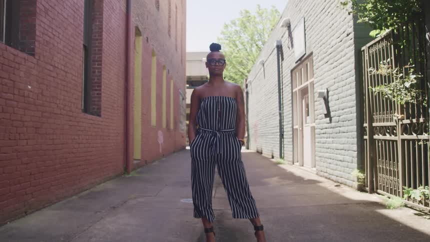 A woman in a strapless jumpsuit and heels stands in an alley posing on a bright sunny day
