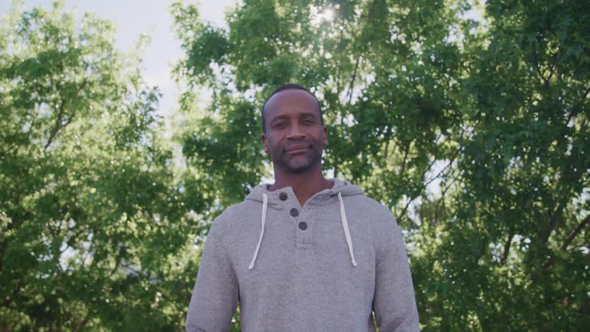 Man wearing hoodie stands outdoors near trees on sunny day smiling slightly