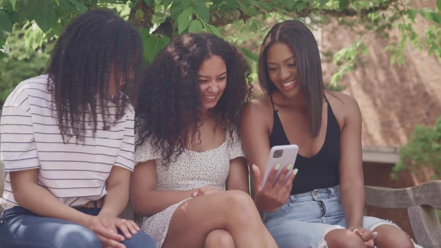 Women with frizzy hair sit in a bench and look at a smartphone laughingly in a garden