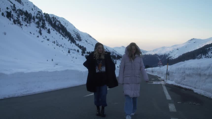 A pair of women in winter coats walk down a road through a snowy mountain valley during the day