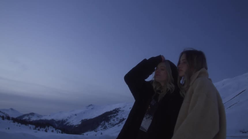Two women with jackets hugging with a mountain range the sky and clouds in the background