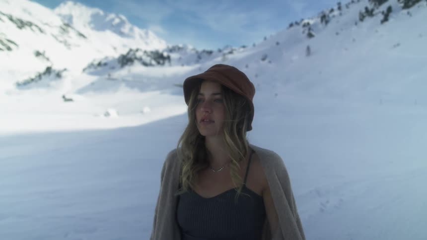 Blonde woman with brown hair and sleeveless shirt with a beige coat stands in front of snowy mountains