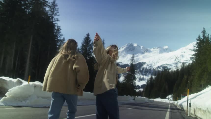 Two girls jumps up and down together on a road surrounded by winter snow and trees