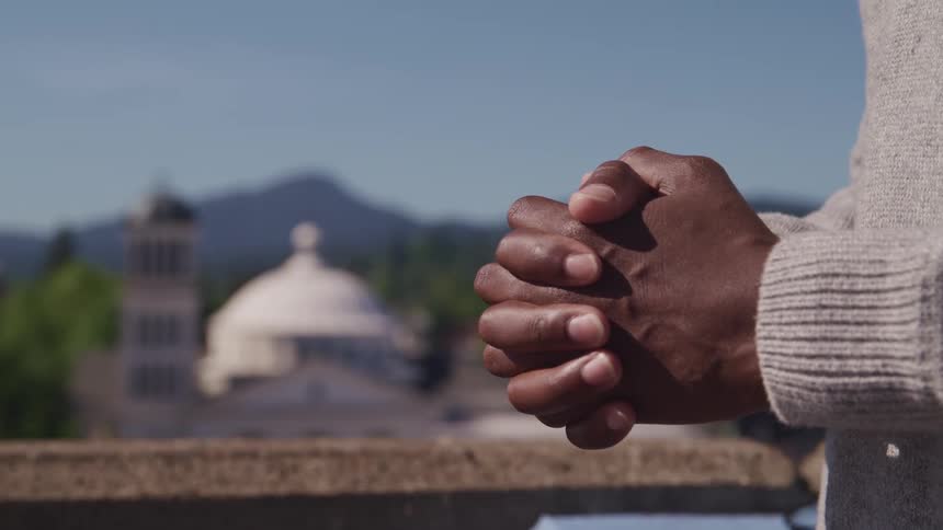 A man with a sweater praying with his hands folded with a church trees the sky and clouds in the background