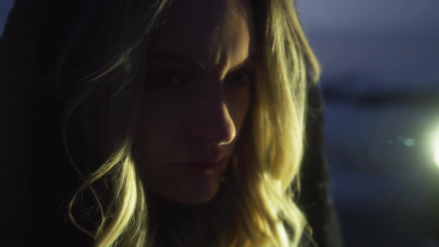 A blonde woman standing outside at night with a bright light in the background