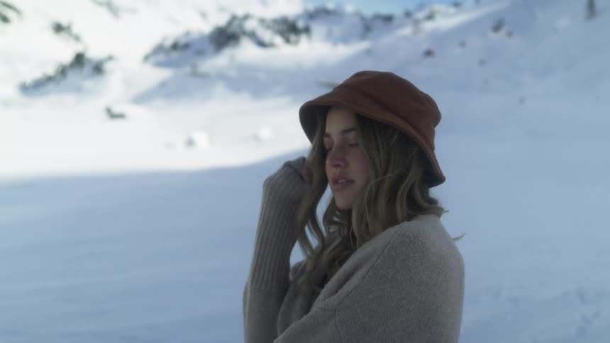 A woman with a sweater and a hat standing with a mountain range the sky and clouds in the background