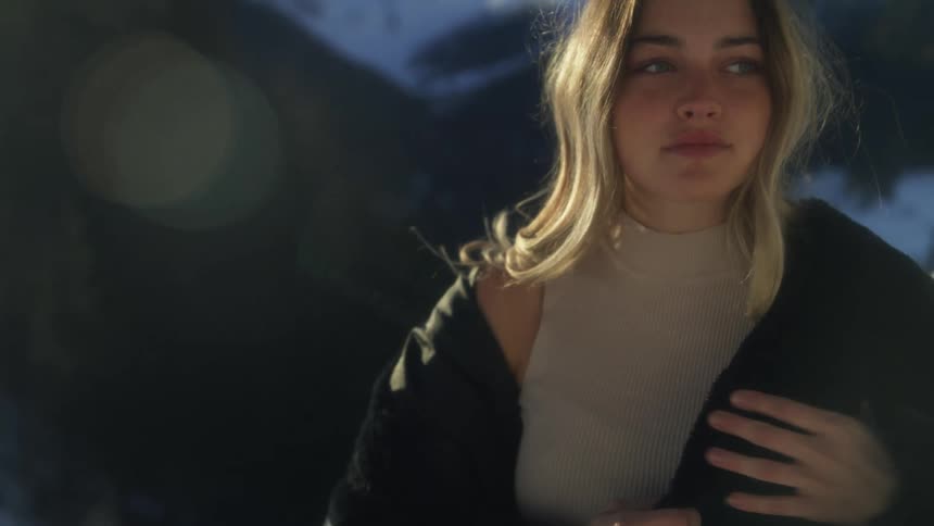 Blonde woman wears a black coat and stands against a background of mountains