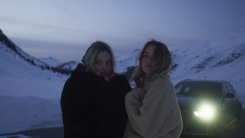 Two women with coats with a vehicle a mountain range the sky and clouds in the background