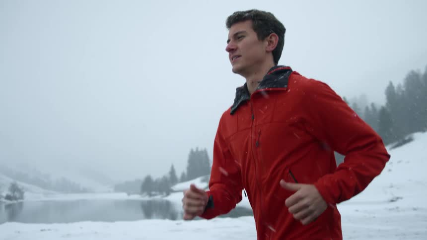 A man jogging while it is snowing with a lake trees the sky and clouds in the background