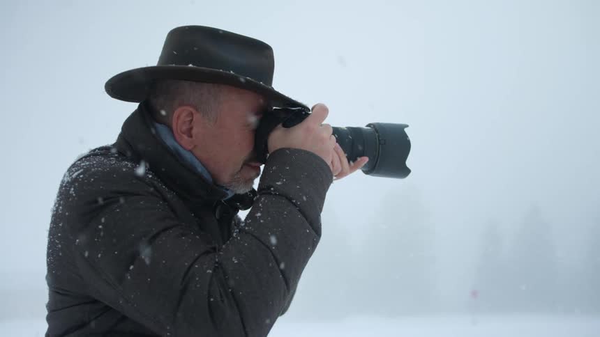A man standing outside in the snow aims his camera to take a photograph