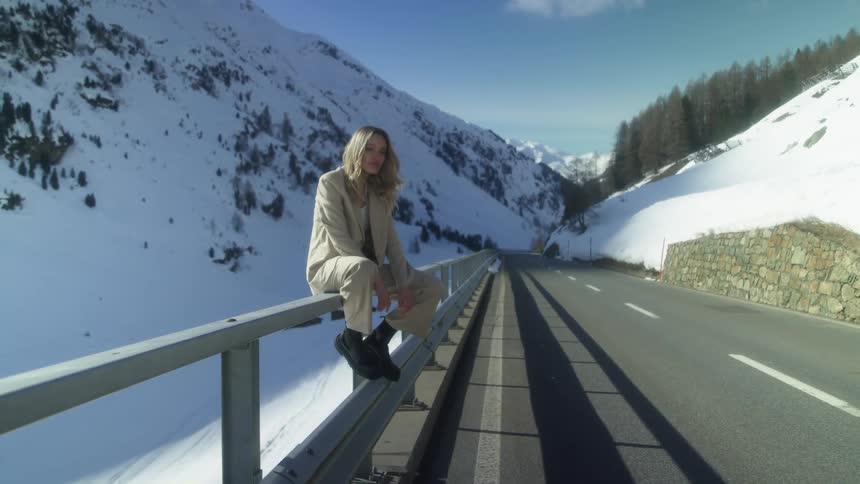 A woman sitting on railing by a road with a mountain range trees the sky and clouds in the background