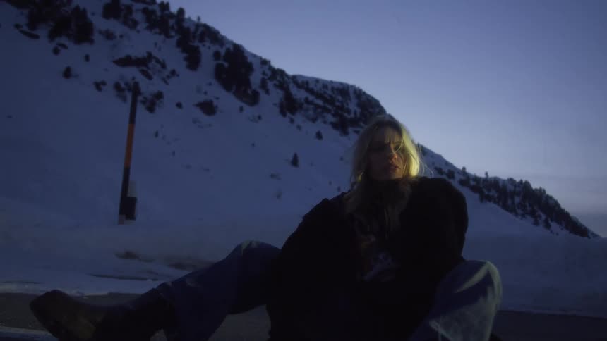 Blonde woman sitting in a road in the snow covered mountains at sunset